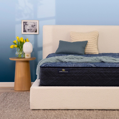 Close-up of a Serta Perfect Sleeper mattress featuring a navy quilted side with a brand label and a dark blue sunburst pattern on top, styled in a cream boucle bed frame with decorative pillows.