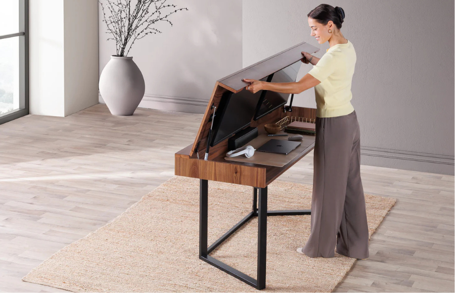 A woman in a yellow shirt and brown trousers stands at a modern wooden desk with black metal legs. She is lifting the hinged desktop panel, which has two computer monitors attached to its underside. The desk surface underneath holds a laptop, a notebook, and headphones. In the background, there is a large white vase with bare branches on a light wooden floor.
