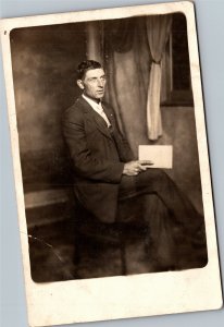 RPPC Man holding book seated in chair by window