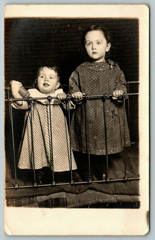 RPPC Toddler w/Bottle and Big Sister Stand Behind Metal Bed Railingc1910 