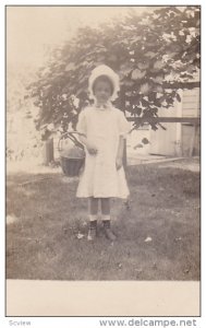 RP; Little girl wearing bonnet, holding wicker basket, standing in yard, 1910...