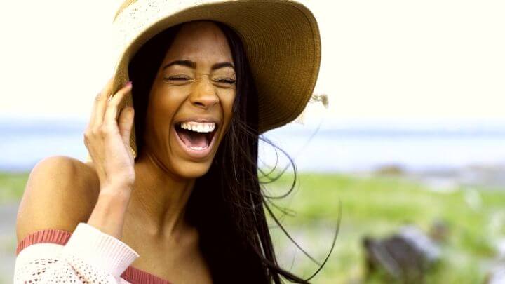 African American woman at the beach holding her floppy hat, laughing