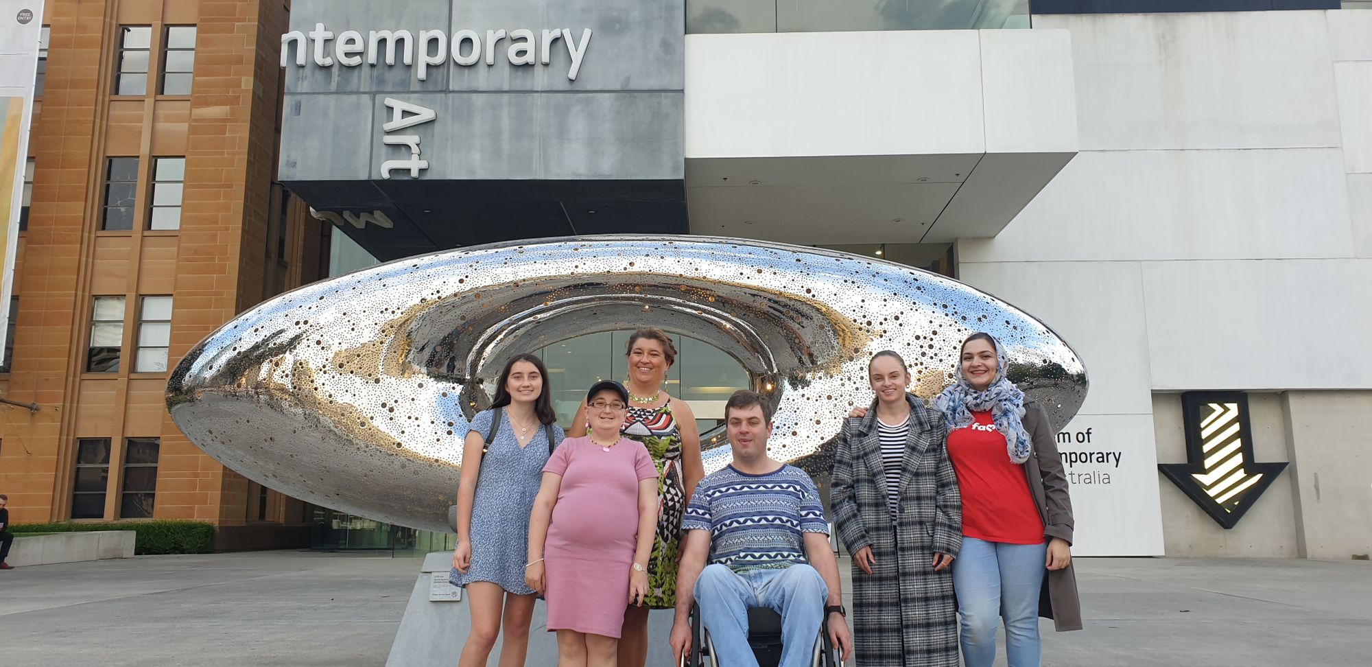 A group of six smiling people, including one person in a wheelchair, stand in front of a silver sculpture. The MCA building is in the background.