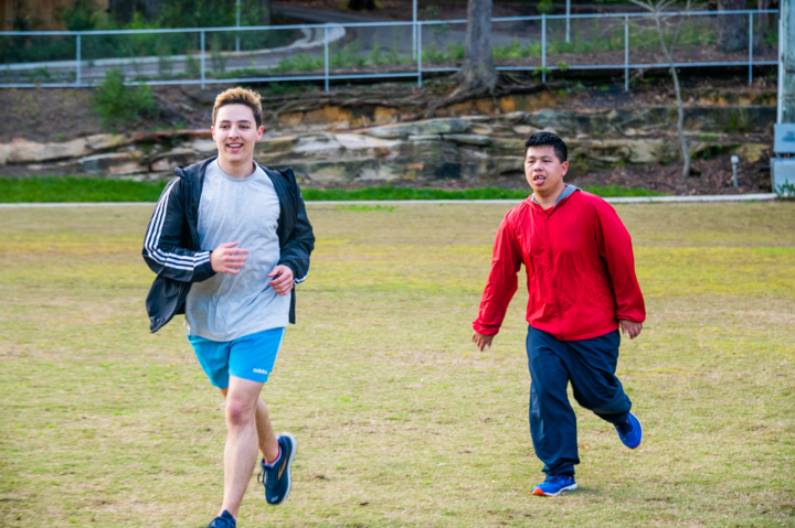 A man in a grey shirt and blue shorts is running on an oval, followed by a man in a red jacket and navy tracksuit pants 