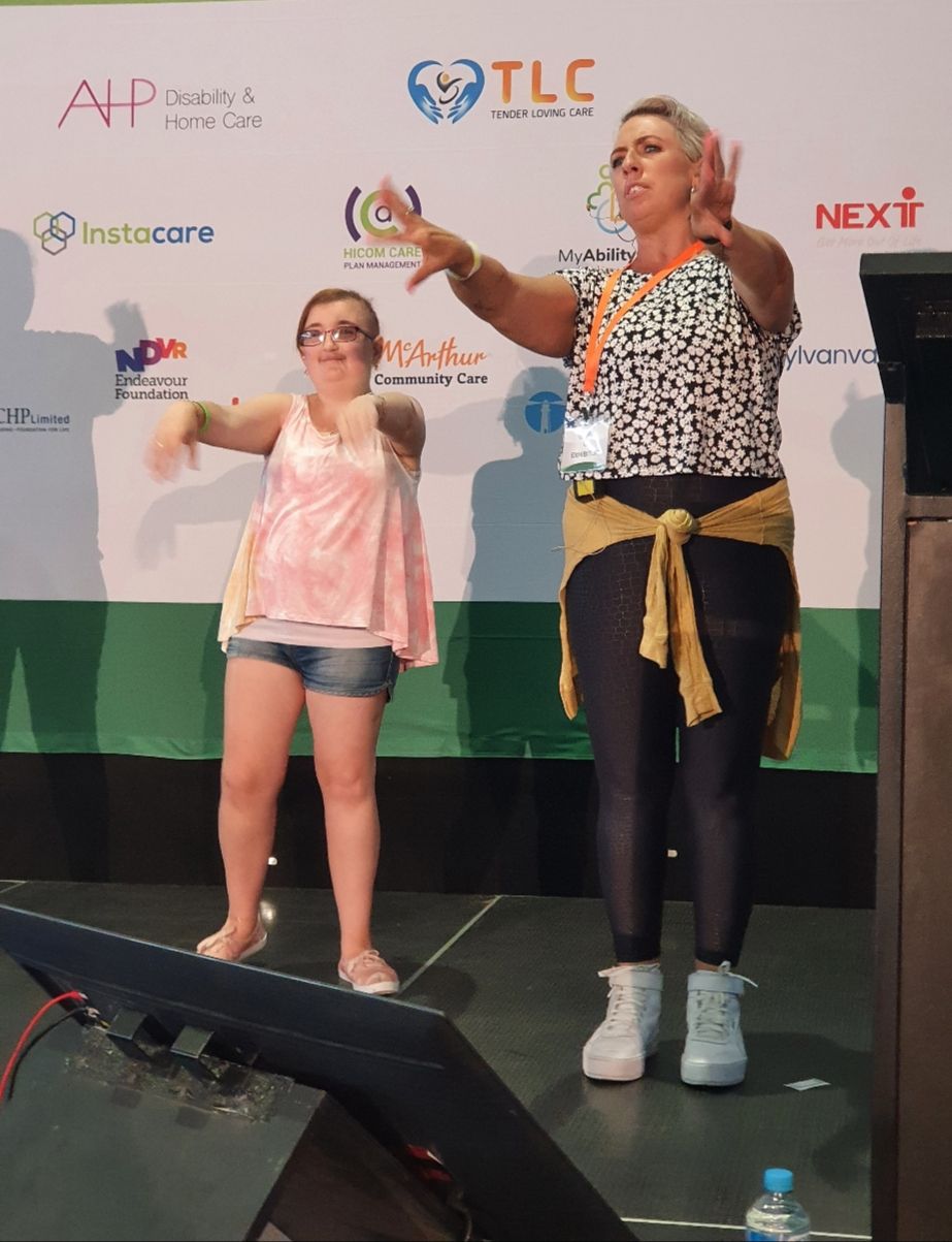 Two women stand in front of a poster with their arms stretched in front of them. The one on the left wears a pink shirt and blue shorts. The woman on the right wears a black and white shirt with black leggings.