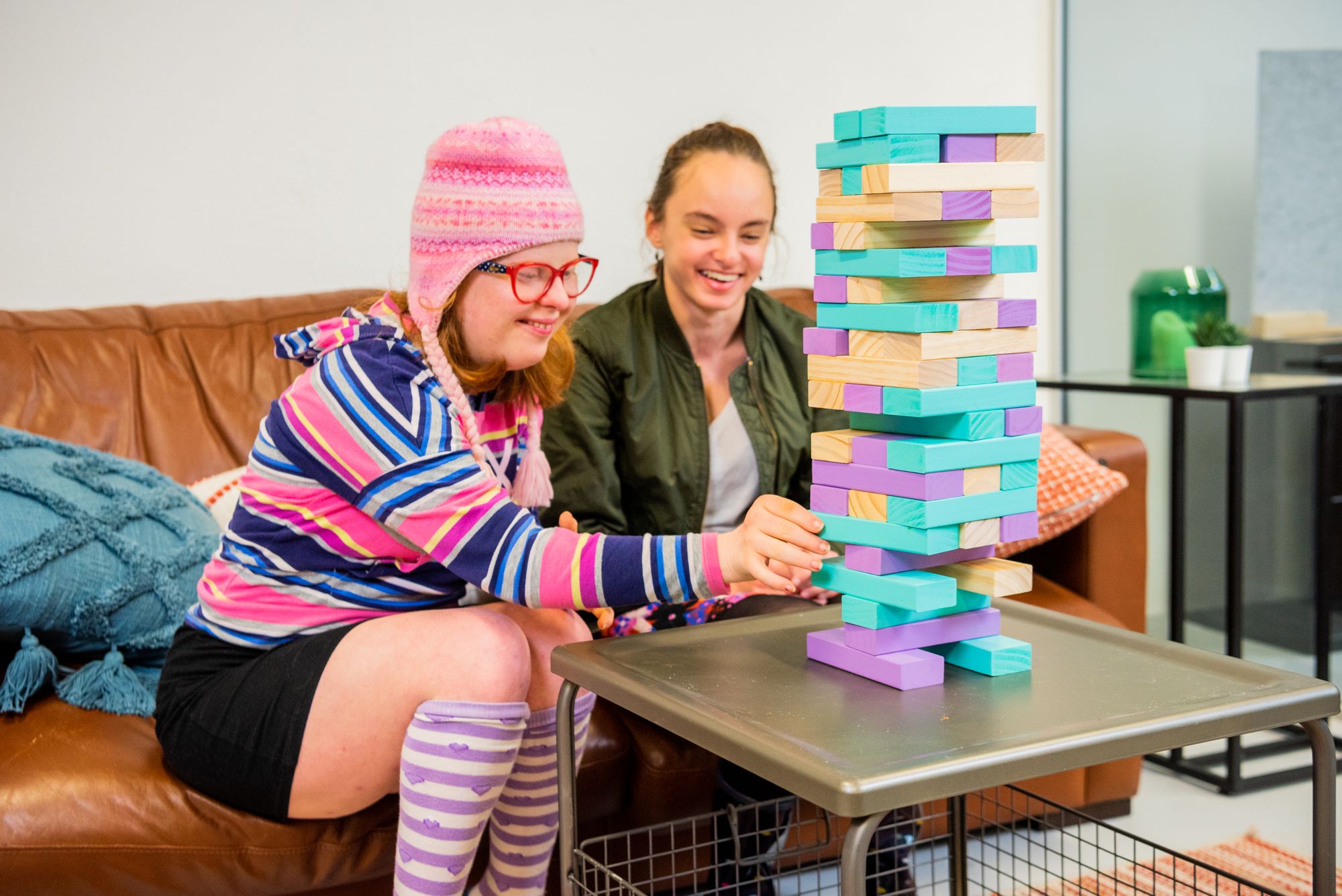 Two women sit on a couch playing Jenga with colourful blocks