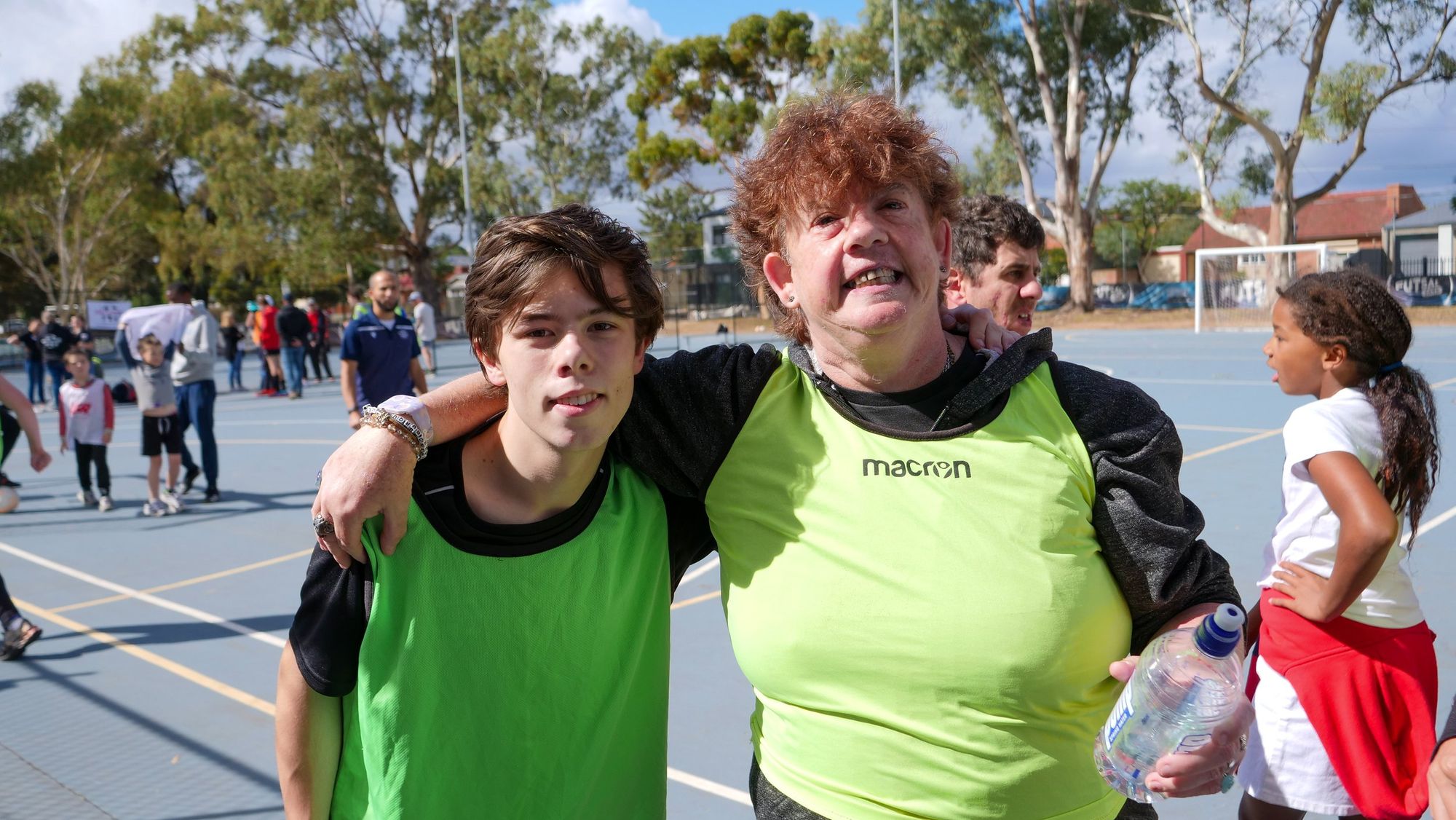 A woman in a yellow sports bib stands on a court with her arm around a boy in a green bib