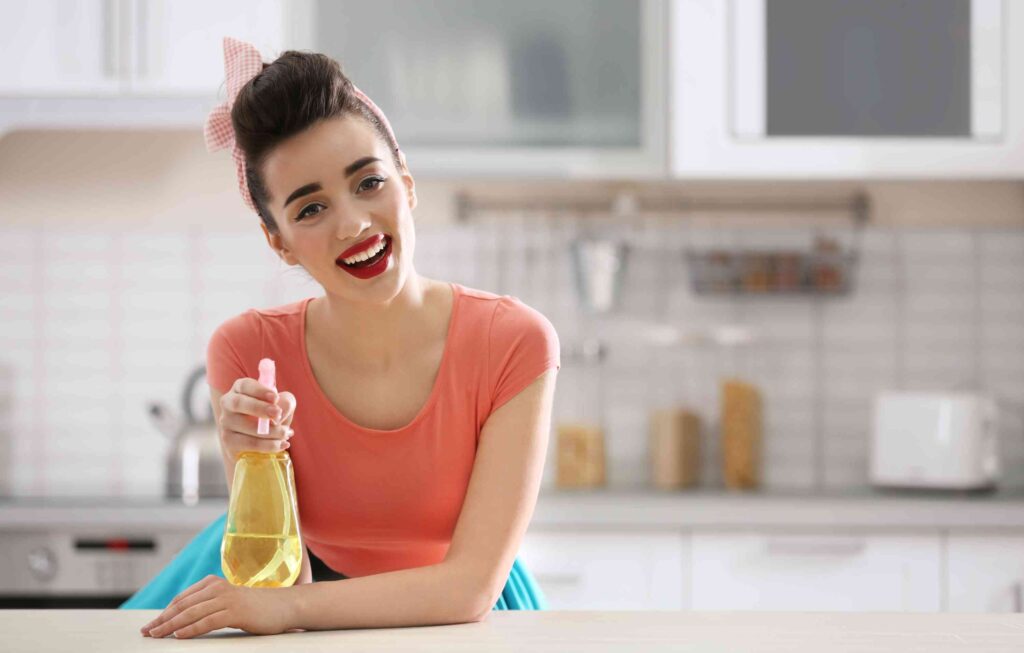 woman holding spray bottle in front, smiling. Kitchen background, grayed out in the back