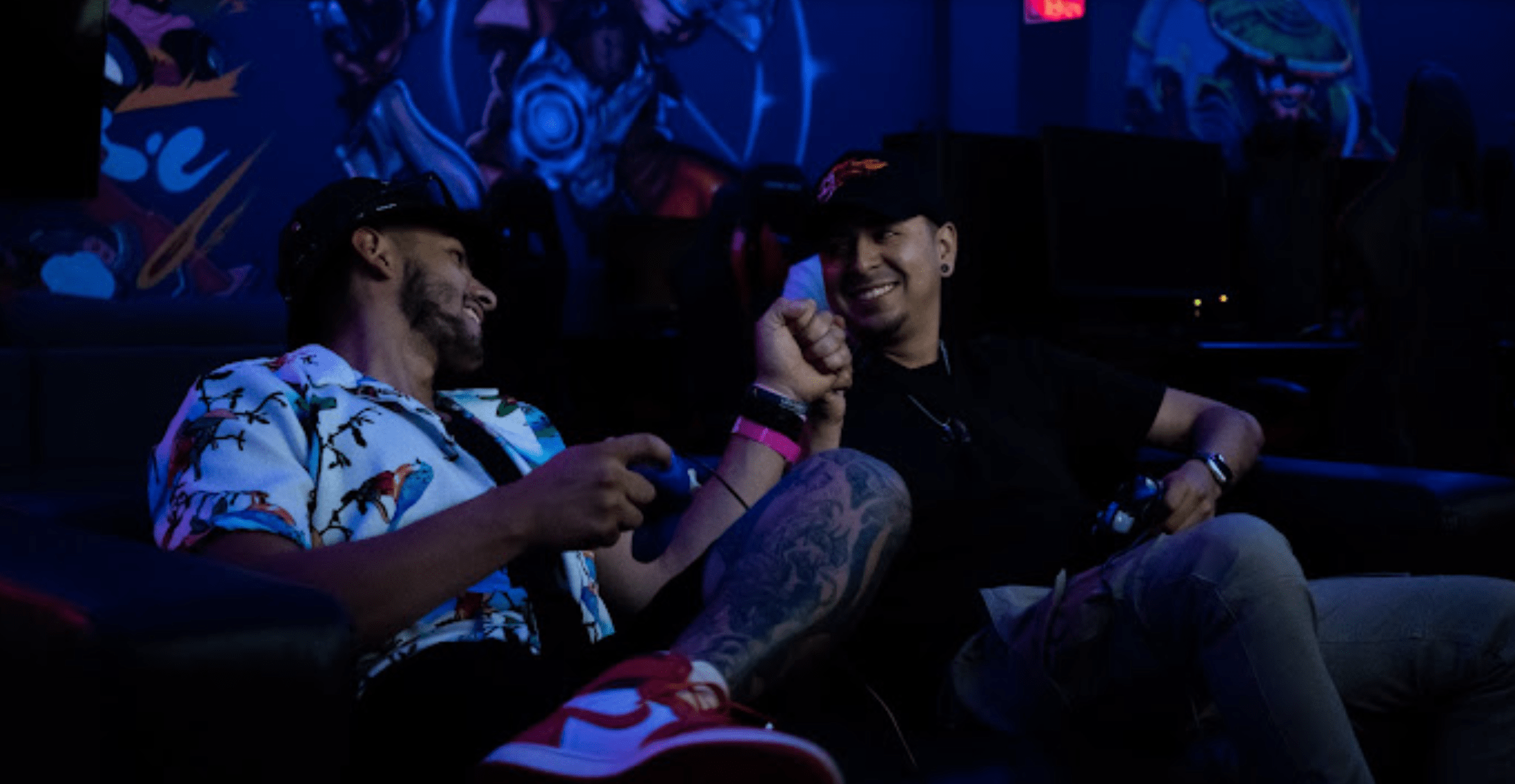 Two men sitting on a couch playing video games in a dimly lit room with blue lighting and game art on the wall
