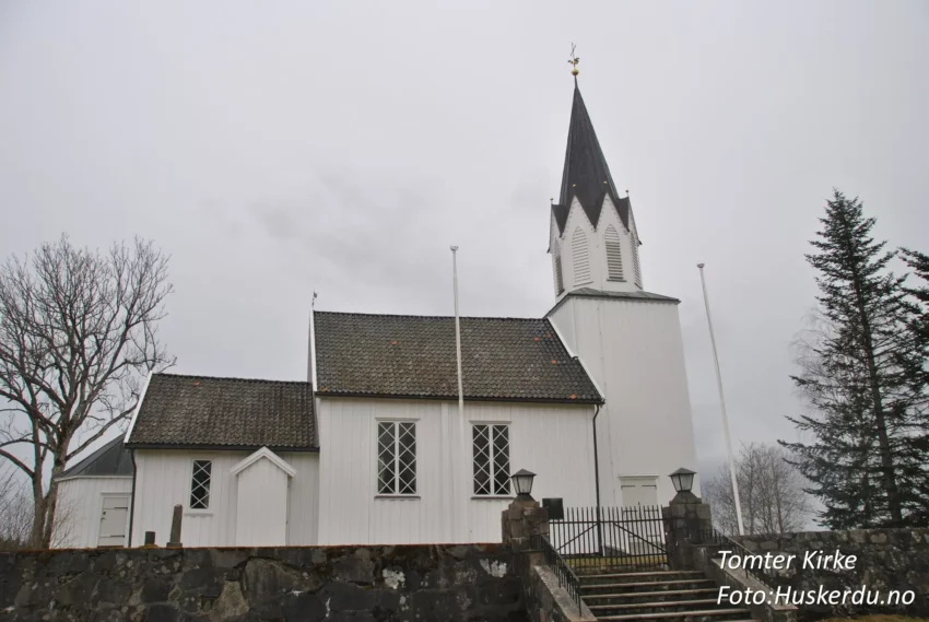 Tomter kirke, en tradisjonell langkirke i Indre Østfold, Østfold fylke, er bygget i tømmer og har en rik historie som strekker seg tilbake til 1600-tallet. Kirken er kjent for sin nygotiske altertavle fra 1880 og vakre kirkegård, som reflekterer områdets kulturarv. Foto: Huskerdu.no