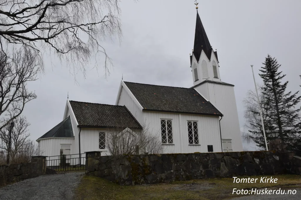 Tomter kirke, en tradisjonell langkirke i Indre Østfold, Østfold fylke, er bygget i tømmer og har en rik historie som strekker seg tilbake til 1600-tallet. Kirken er kjent for sin nygotiske altertavle fra 1880 og vakre kirkegård, som reflekterer områdets kulturarv. Foto: Huskerdu.no