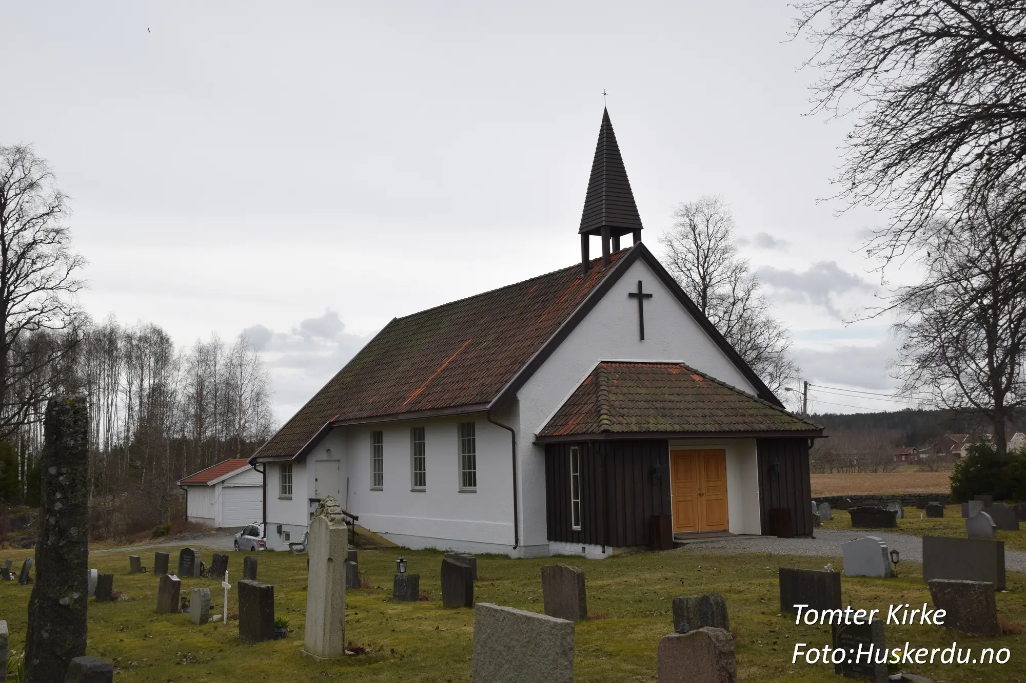 Tomter kirke, en tradisjonell langkirke i Indre Østfold, Østfold fylke, er bygget i tømmer og har en rik historie som strekker seg tilbake til 1600-tallet. Kirken er kjent for sin nygotiske altertavle fra 1880 og vakre kirkegård, som reflekterer områdets kulturarv. Foto: Huskerdu.no