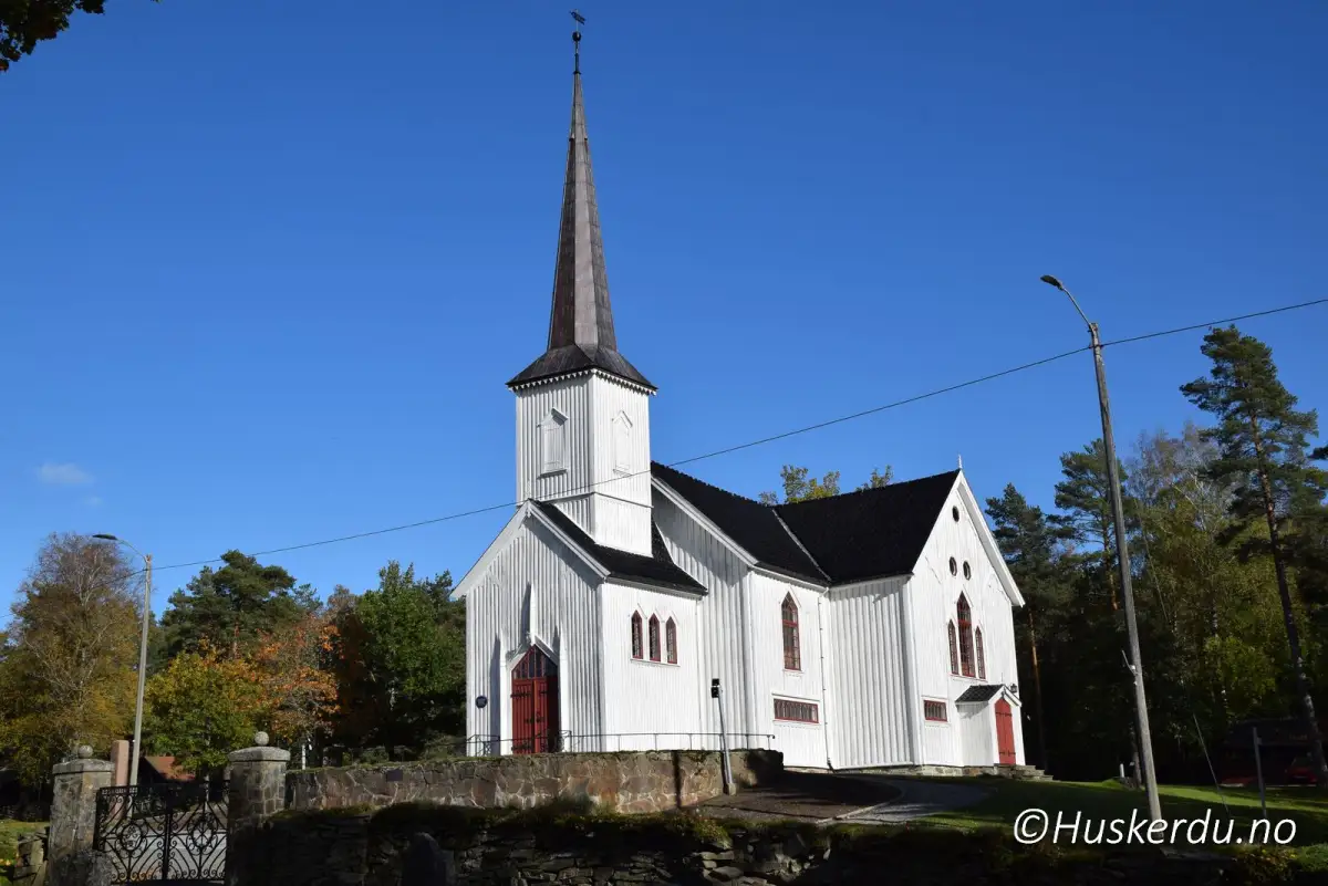 Båstad kirke blant høstfarger 🍂⛪ Den hvitmalte trekirken reiser seg stille over en lav steinmur, omgitt av gressplener og trær med fargesprakende løv. Dette bildet av Båstad kirke i Indre Østfold er tatt en klar høstdag, med blå himmel og lune farger rundt. Kirken ligger rett ved veien og er lett å finne for reisende som vil se nærmere på lokale bygdetun og eldre kirkebygg. Det røde kirketårnet gir et diskret blikkfang i landskapet, og området rundt gir plass til både stillhet og foto. Foto: © Huskerdu.no bastad-kirke-indre-ostfold-hoststemning Båstad kirke – Høstfarget landskap rundt hvit trekirke i Indre Østfold Båstad kirke, Bastad kirke, Båstad Church, Baastad kirke, Indre Østfold, trekirke Østfold, norsk landsbykirke, høst