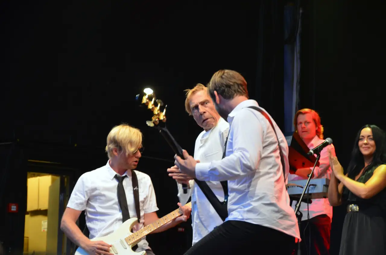 Jahn Teigen på scenen 🎶 Jahn Teigen i energisk samspill med musikere under en konsert. Kledd i hvitt midt på scenen møter han gitaristene tett på, mens bandmedlemmene bak følger opp med rytmer og kor. Den levende scenestilen og kontakten med både publikum og medmusikere gjorde ham til en uforglemmelig artist. Konserter som dette ble minneverdige øyeblikk som løftet norsk musikk ut til et bredt publikum. Foto: Bamble kommune, CC BY-SA 2.0 https://creativecommons.org/licenses/by-sa/2.0 https://commons.wikimedia.org/wiki/File:Jahn_Teigen_(91)_(5982880944).jpg jahn-teigen-konsert-bamble-2011 Jahn Teigen på scenen i Bamble bamble, bamle, Telemark, Telemark fylke, jahn teigen, jan teigen, konsert, norsk musikk Bamble kommune