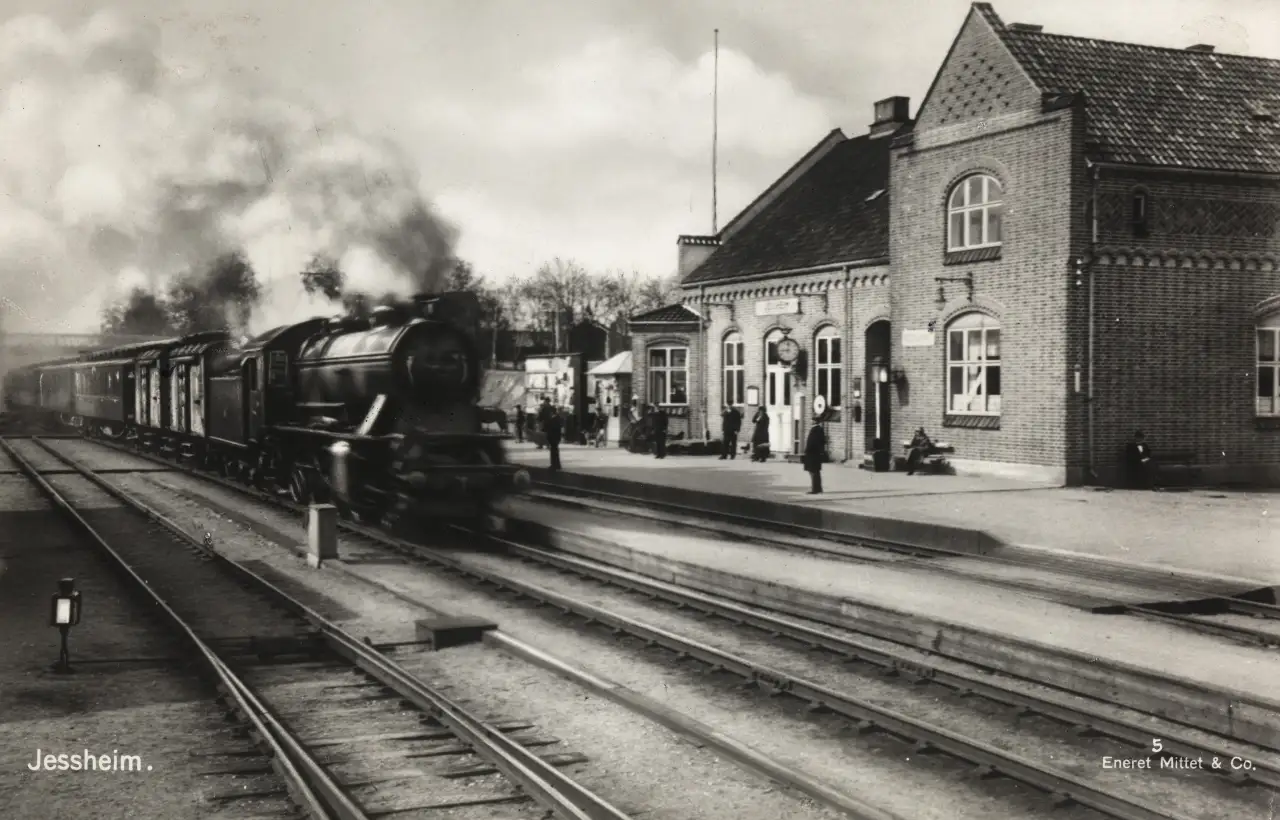 Dampskipstiden på Jessheim 🚂 Et damplokomotiv ankommer Jessheim stasjon tidlig på 1900-tallet, omgitt av reisende og ansatte på perrongen. Den karakteristiske murbygningen fra 1908 danner bakteppet for et glimt inn i en tid da jernbanen var livsnerven på Øvre Romerike. Stasjonen var et knutepunkt for både handel og reiser, og mye av Jessheims utvikling startet nettopp her. Foto: Nasjonalbiblioteket - Public Domain Nasjonalbiblioteket - Public Domain https://commons.wikimedia.org/wiki/File:5._Jessheim_(22666332937).jpg https://upload.wikimedia.org/wikipedia/commons/f/fc/5._Jessheim_%2822666332937%29.jpg dampskipstiden-pa-jessheim-stasjon Jessheim stasjon med damplokomotiv – Historisk togankomst Jessheim, Jessheim stasjon, damplokomotiv, tog Jessheim, jernbanestasjon Jessheim, Øvre Romerike, Ullensaker, Akershus Ullensaker kommune