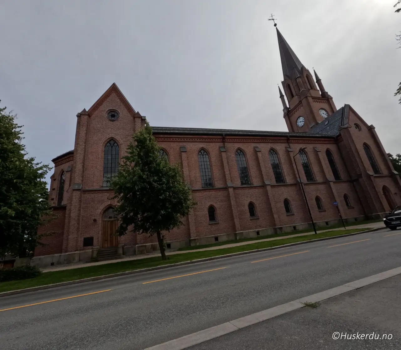 Fredrikstad domkirke i Østfold med høyt tårn og røde teglmurer, omgitt av trær og byliv langs Glomma