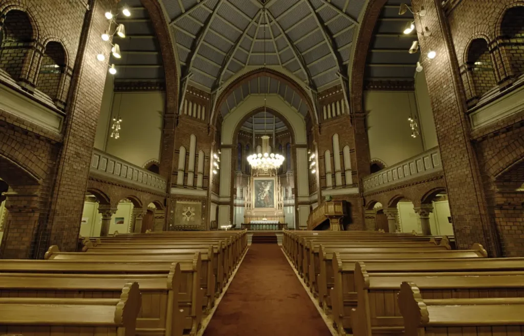 Interiør fra Sagene kirke i Oslo med benker, høyt tak, lysekrone og alter med maleri av Jesu nedtakelse.