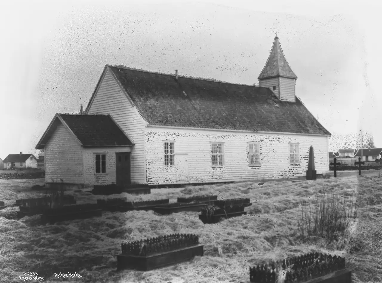 Åkra gamle kirke fotografert i 1925 med kirkegården rundt og bolighus i bakgrunnen.