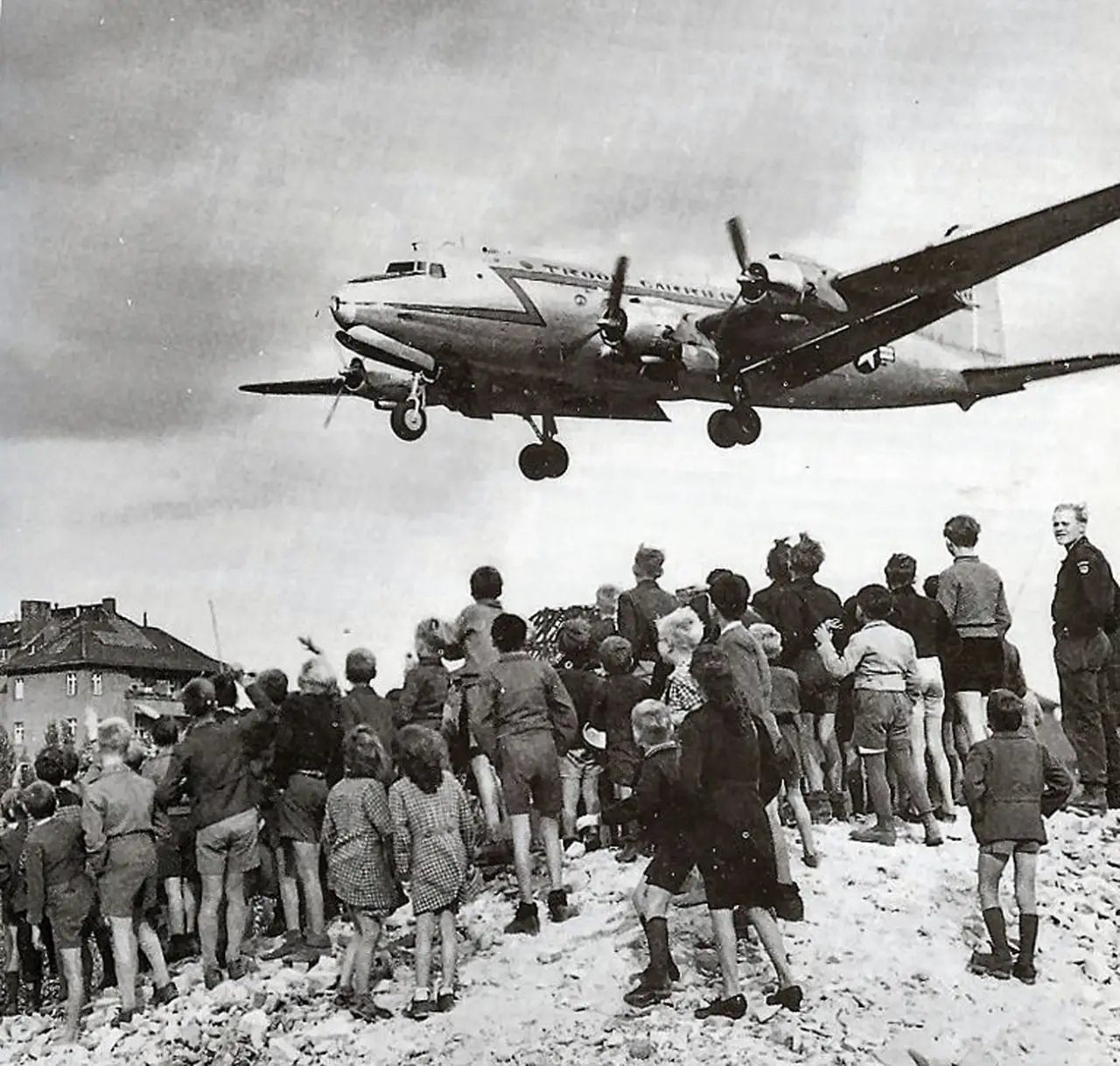 Amerikansk C-54 Skymaster flyr lavt inn mot Tempelhof mens en gruppe barn i West-Berlin står i ruinene og ser opp mot flyet under Berlin-blokaden i 1948.