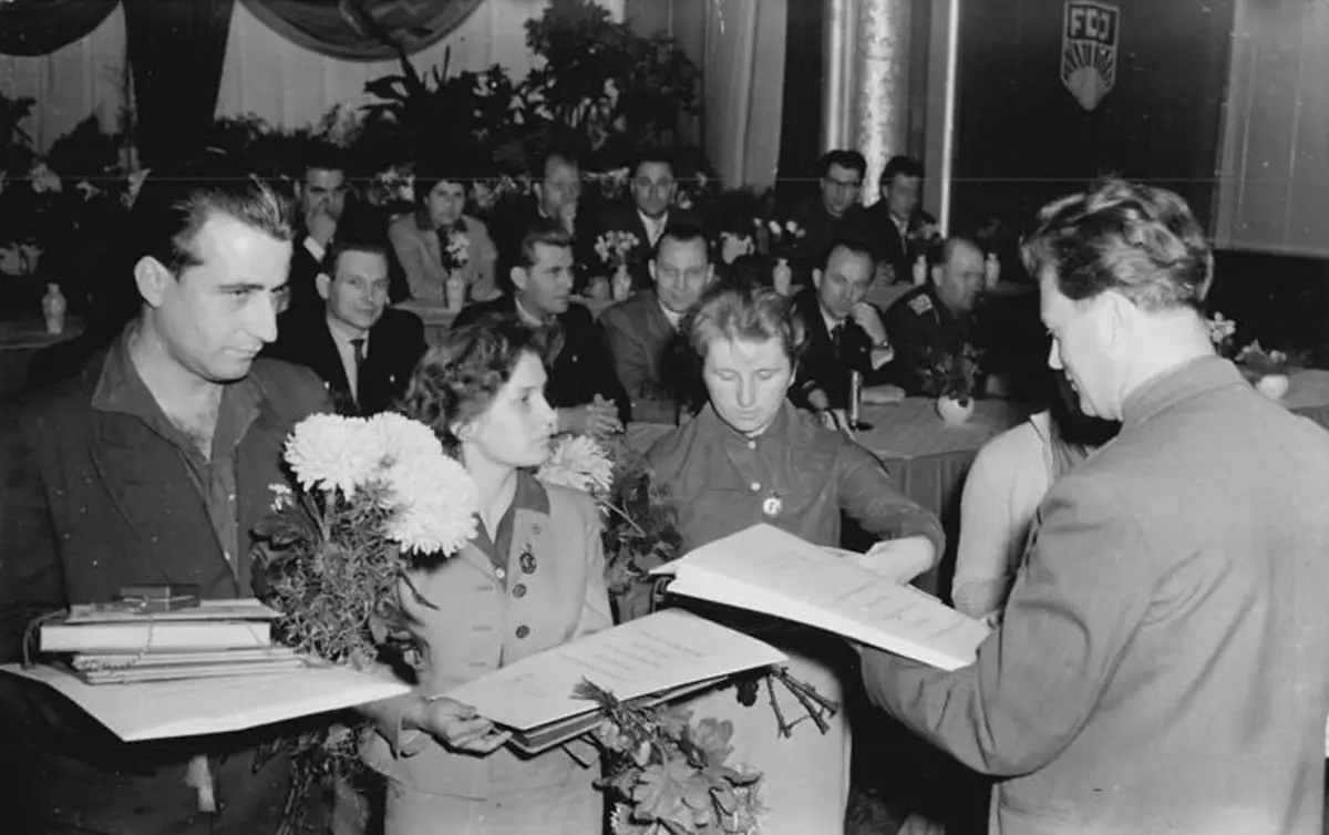 Unge FDJ-medlemmer i DDR får utdelt utmerkelser under en seremoni i 1960, omgitt av partifolk og blomster.