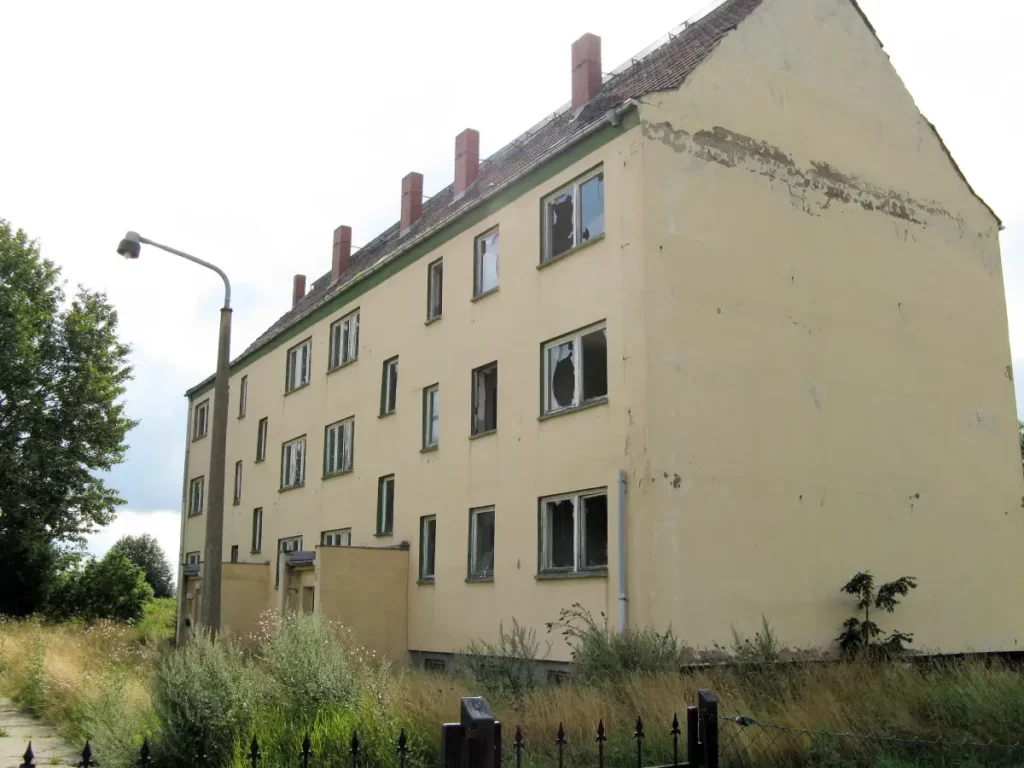 Forlatt og forfallen Plattenbau-bygning med knuste vinduer i Hohen Luckow, omgitt av høyt gress og tegn på fraflytting.