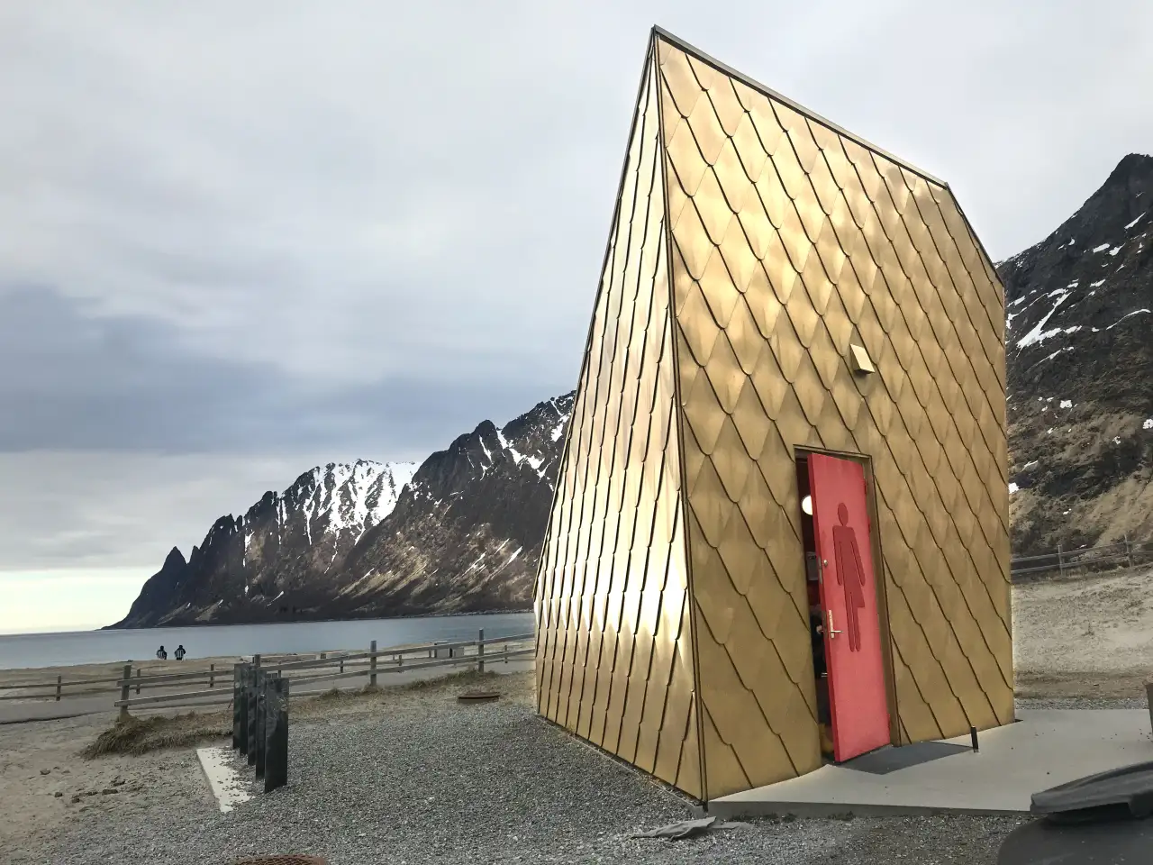 Toalettbygget ved Ersfjord med gullfargede flater og fjellene rundt fjorden i bakgrunnen på Senja.