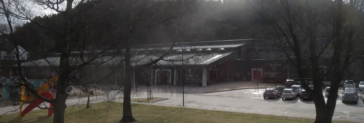 Idda Arena sett fra parkområdet med store trær i forgrunnen og parkeringsplassen ved inngangen.