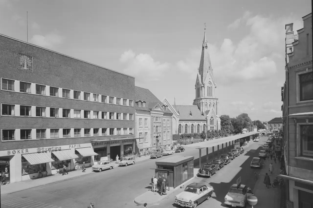 Kristiansand sentrum i 1962 med Domkirken og forretningsbebyggelse langs Henrik Wergelands gate, sett på en travel dag med biler og folk i sentrum.