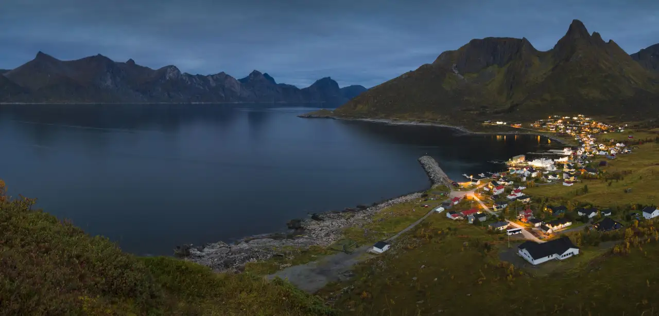 Mefjordvær sett om kvelden med belyste hus, molo og fjellrekke på motsatt side av fjorden.