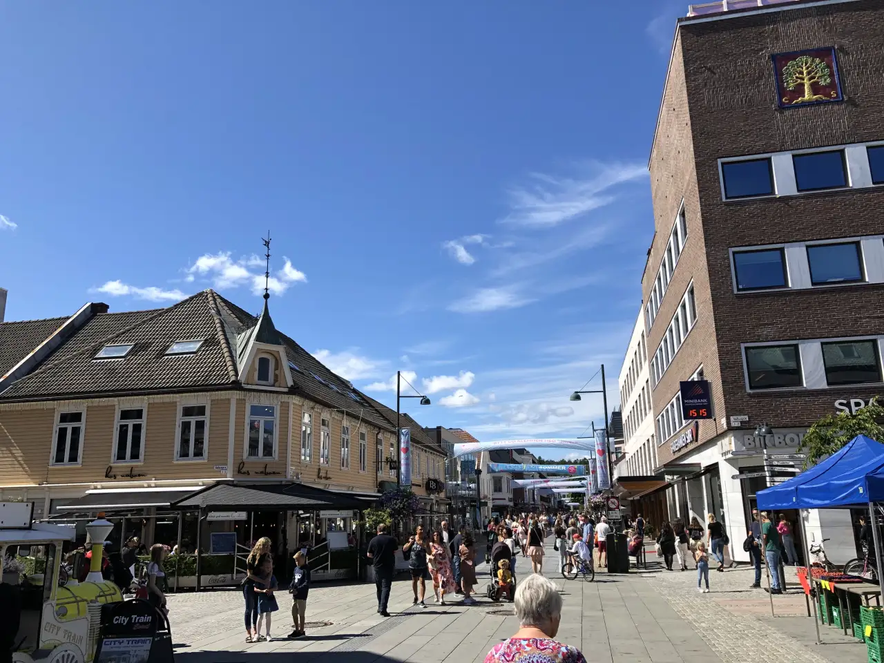 Markens gate i Kristiansand med folk som går langs handlegaten på en solrik dag omgitt av trehus og nyere bygg.