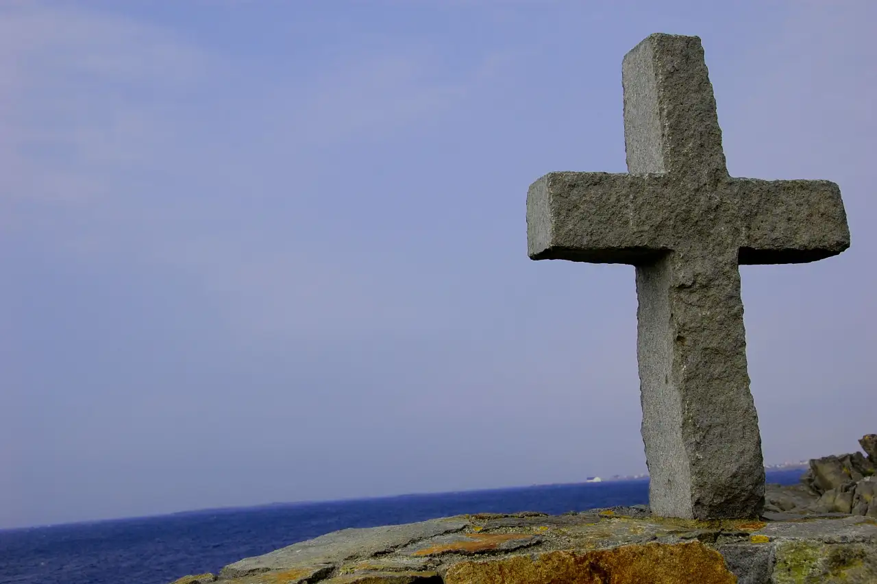 Korsmonument i stein på en liten høyde ved kysten på Ferkingstad med havet i bakgrunnen.