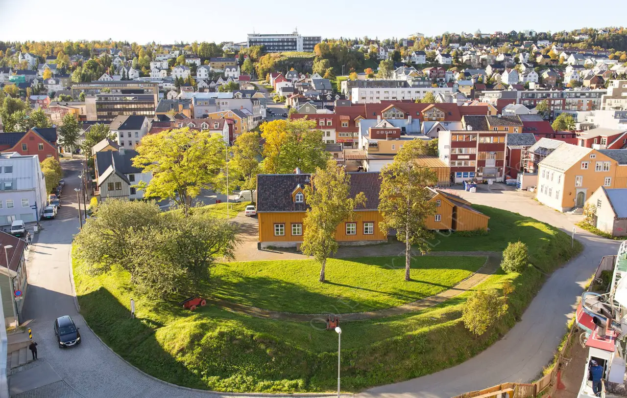 Skansen i Tromsø med gule trebygninger og grønn voll omgitt av sentrumshus.