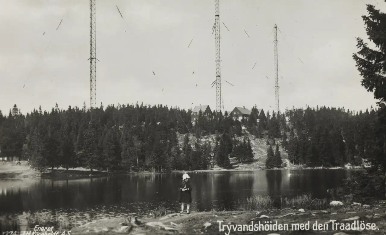 Tryvannshøgda med radiosender og høye master sett fra vannkanten omgitt av skog.