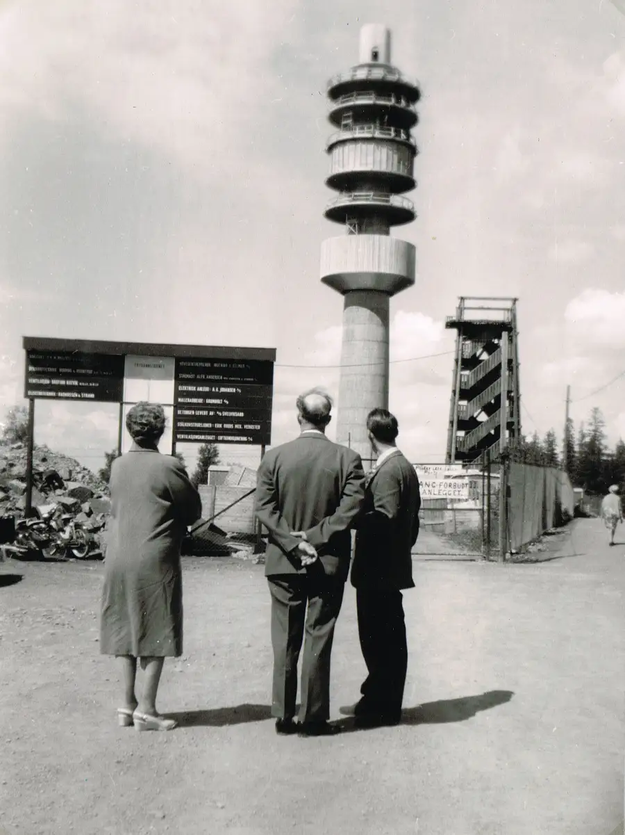 Tryvannstårnet under bygging i 1961 med gammelt tretårn ved siden av og tre personer som ser på.