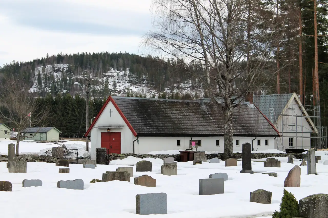 Tuft gravkapell i snødekt vinterlandskap med gravsteiner og skogkledde åser i bakgrunnen.