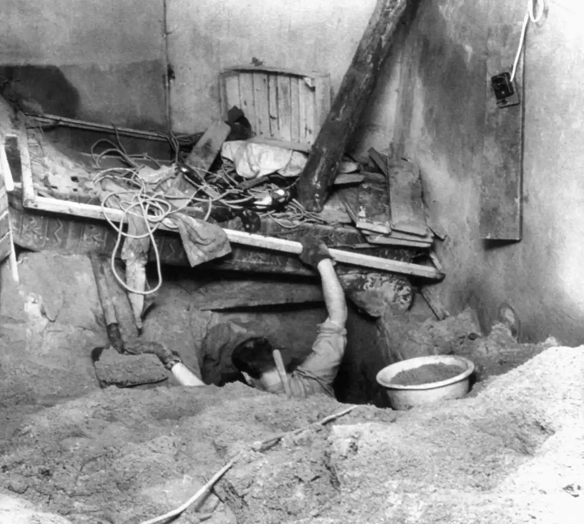 En østtysk flyktning graver en tunnel under Berlinmuren fra en kjeller i Berlin på 1960-tallet, omgitt av jord, enkle verktøy og støttende trebjelker.