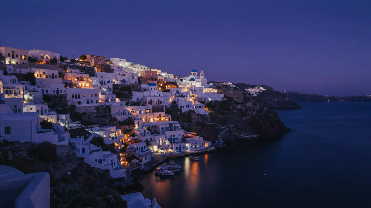 A travel photograph of Santorini at blue hour, white-washed cliffside houses with blue domes, string lights, calm Aegean sea, warm windows glowing, postcard quality