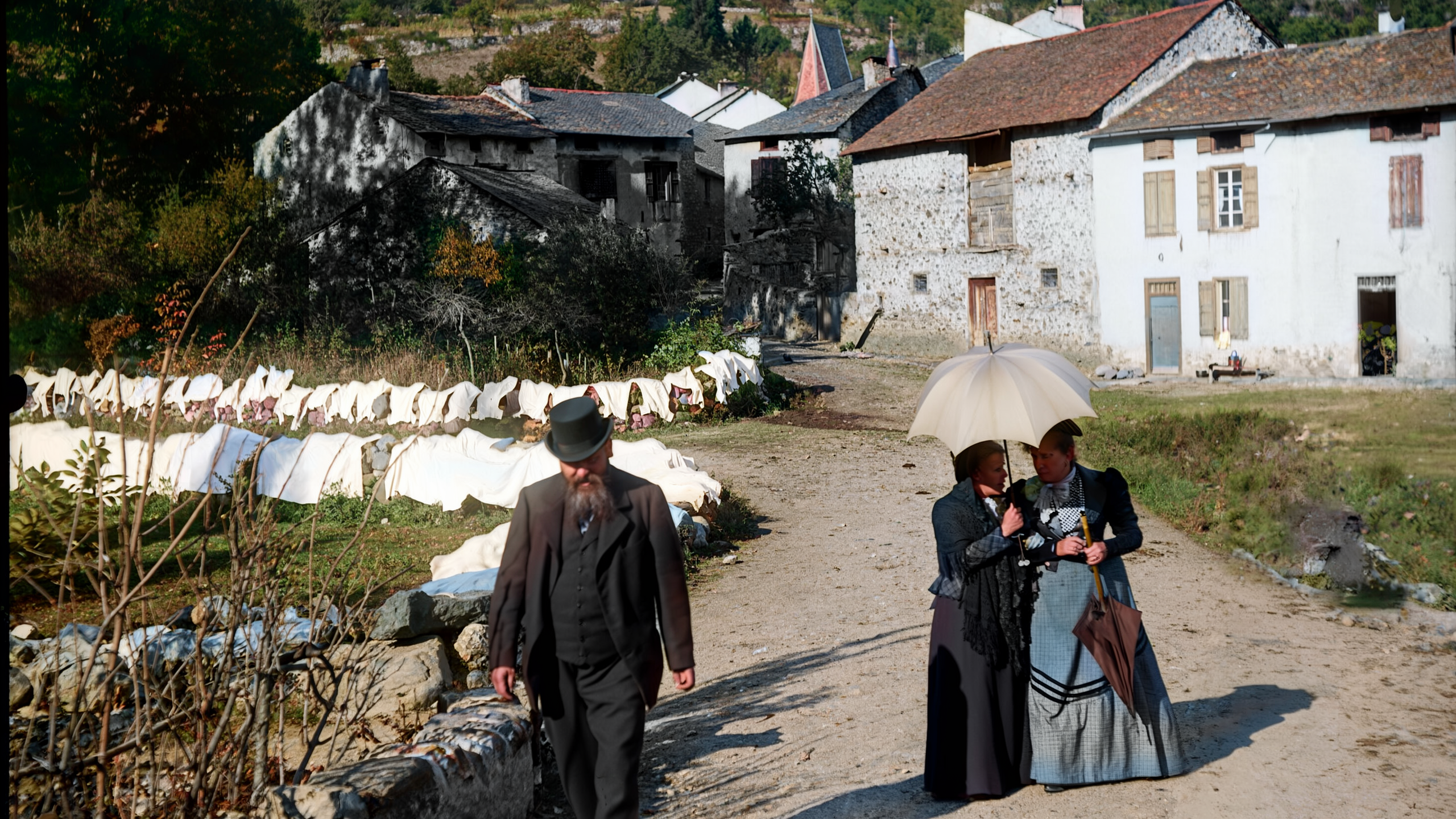 A bearded man in a bowler hat walks along a village path while two wom