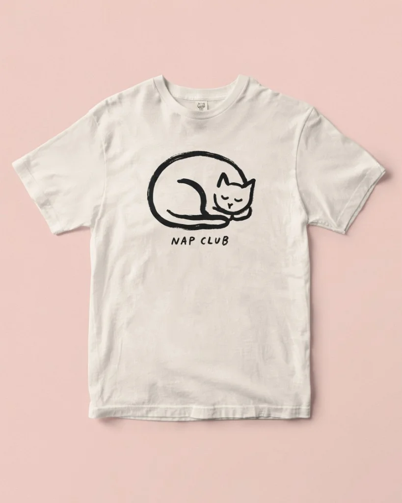 A white t-shirt with a sleeping cat illustration and "NAP CLUB" text on a pink background