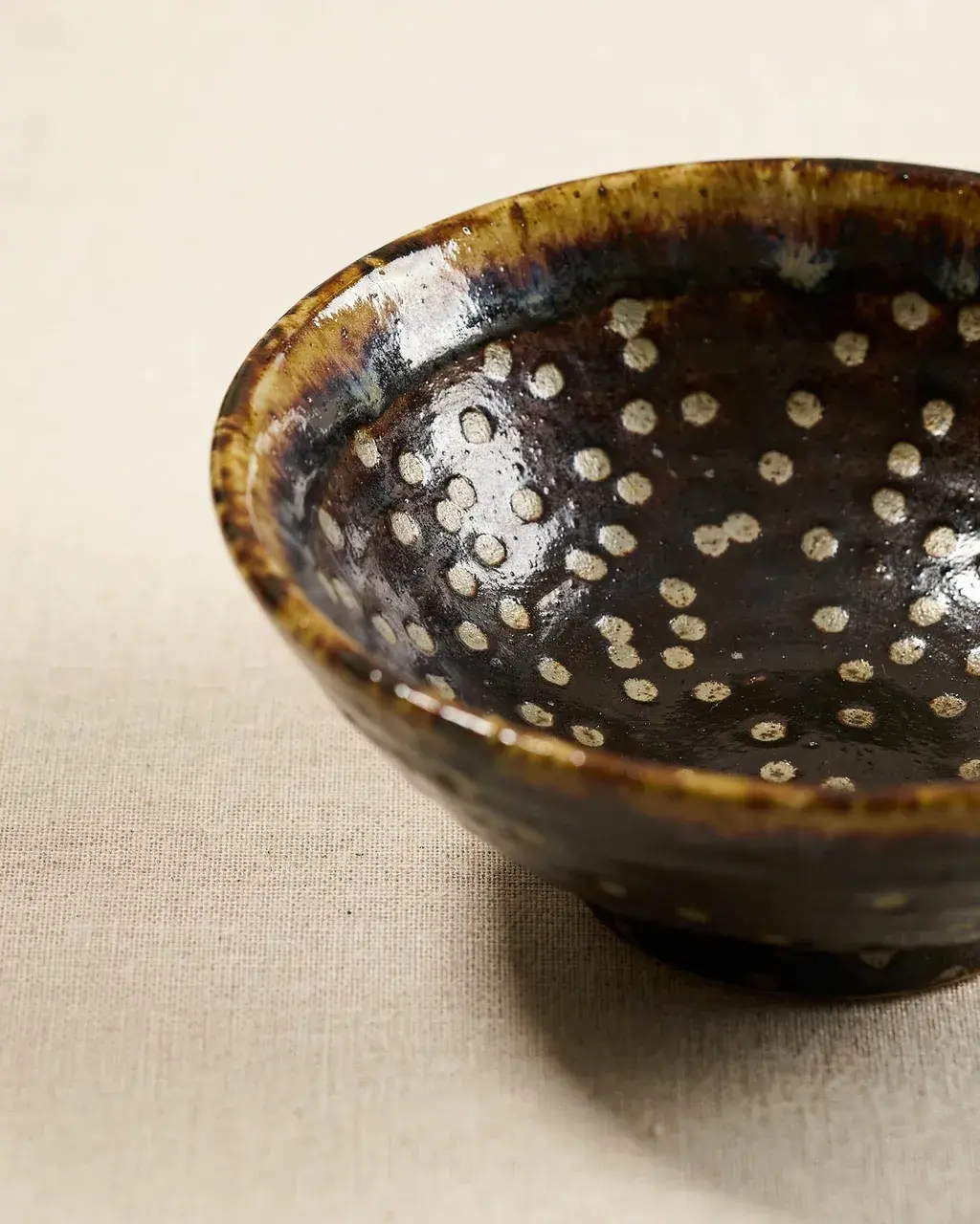 Song dynasty tenmoku tea bowl with oil-spot glaze, iridescent silver-gold spots on deep iron-black, photographed against undyed linen in warm studio light.