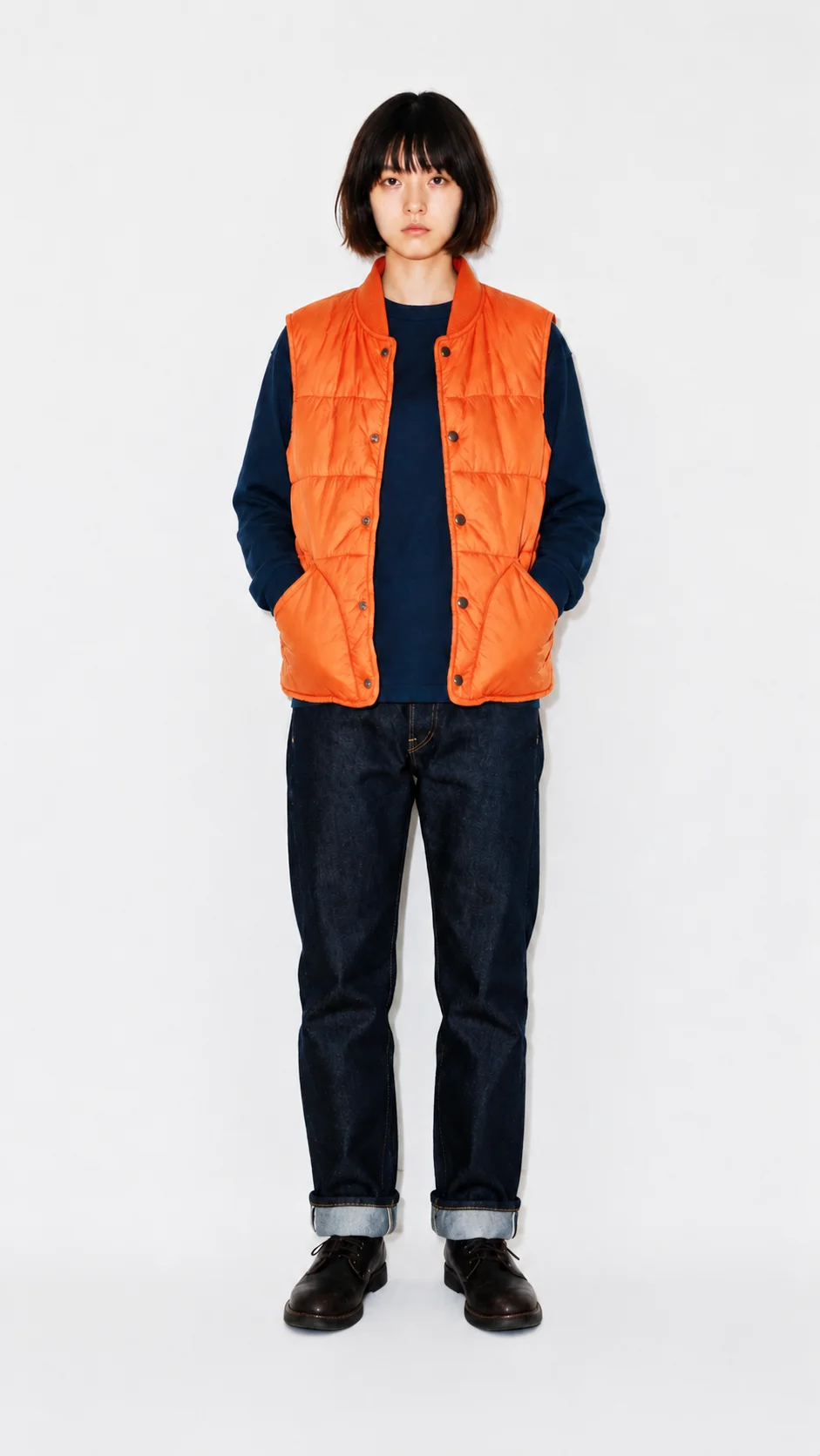 Model in an orange quilted vest, navy tee, and dark denim, photographed in a studio