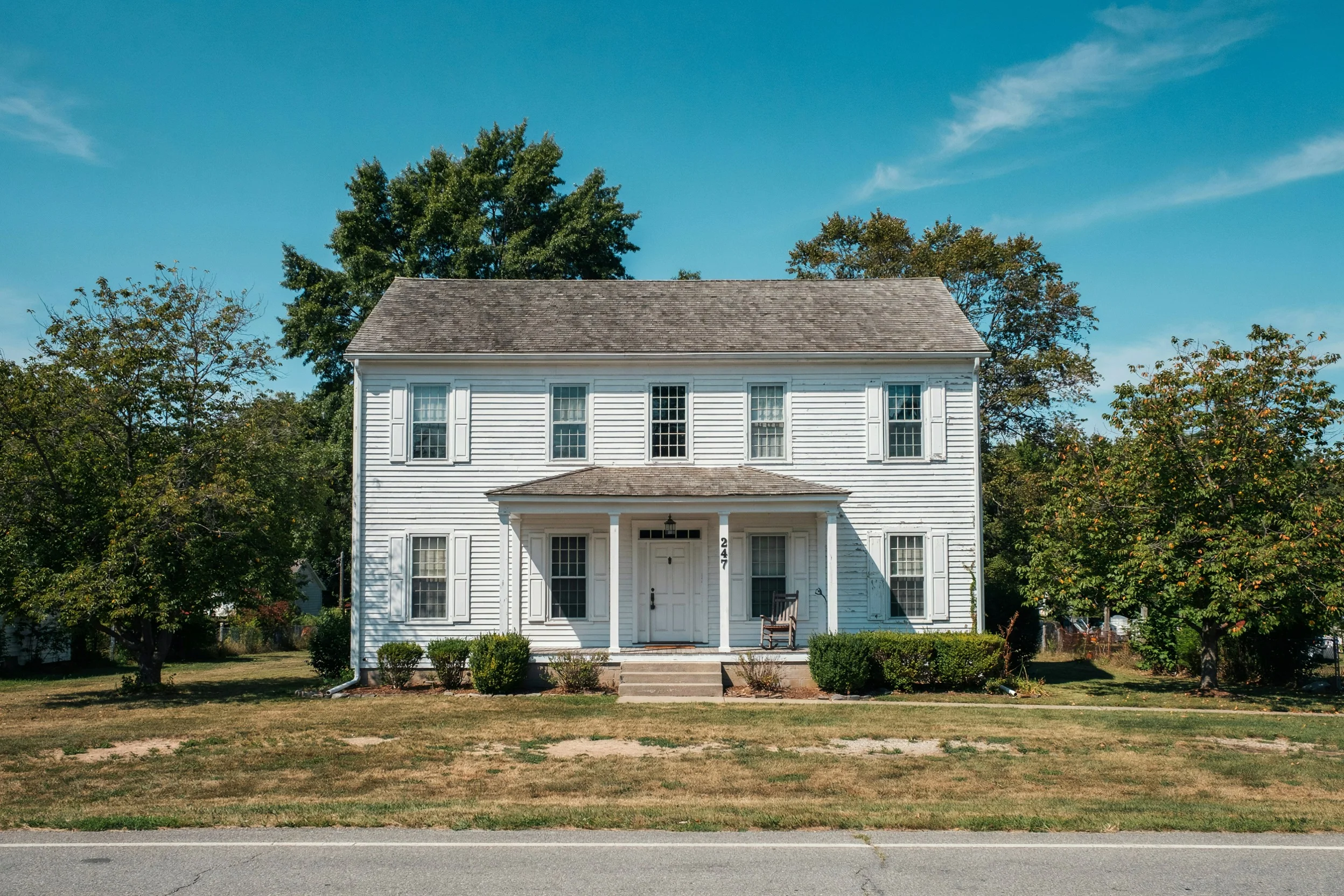 A white clapboard farmhouse with a patchy, dry yellow front lawn