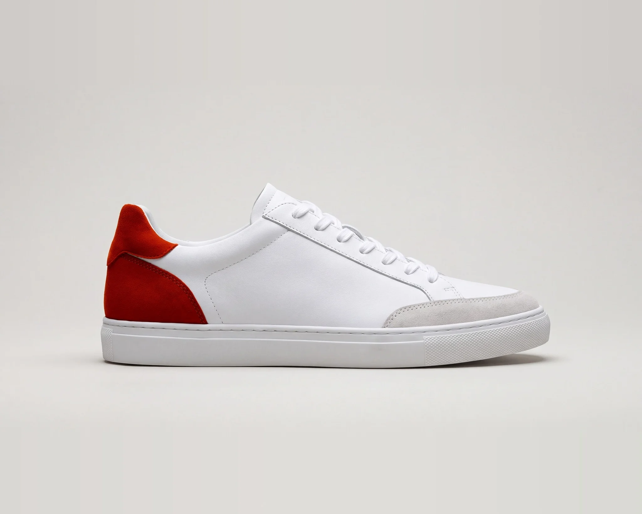 The starting image: a white leather low-top sneaker in side view, toe pointing right, on a light gray background. It has a burnt orange suede heel panel, a light gray suede toe cap, white laces, and a white rubber sole.
