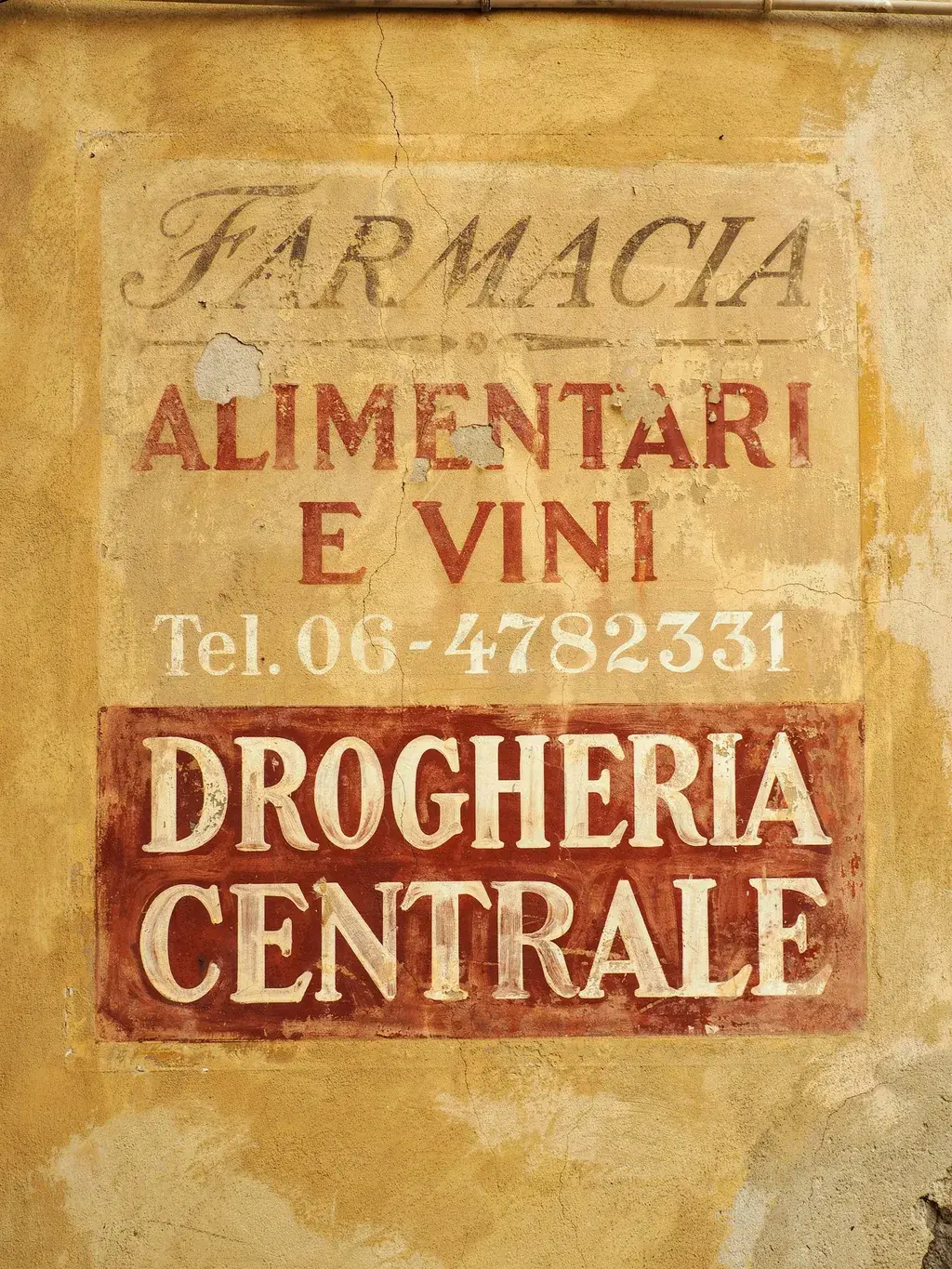 Southern European ochre plaster wall with layered decades of hand-painted signage: FARMACIA faded beneath ALIMENTARI E VINI, phone number, and a bold DROGHERIA CENTRALE on terracotta-red.