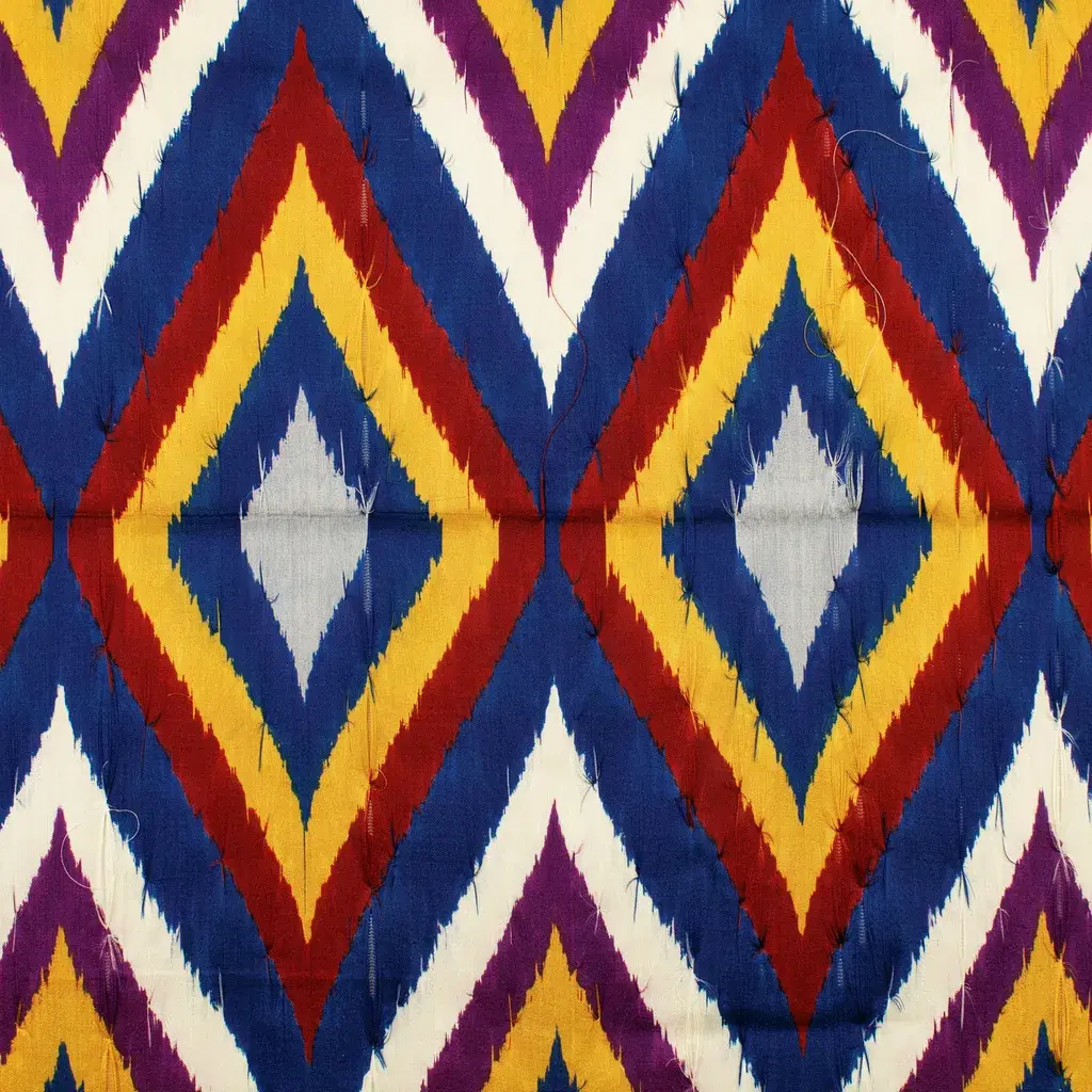 Extreme macro of hand-loomed Central Asian silk ikat: geometric lozenges and chevrons in deep cobalt, cinnabar red, and saffron, individual silk threads visible.