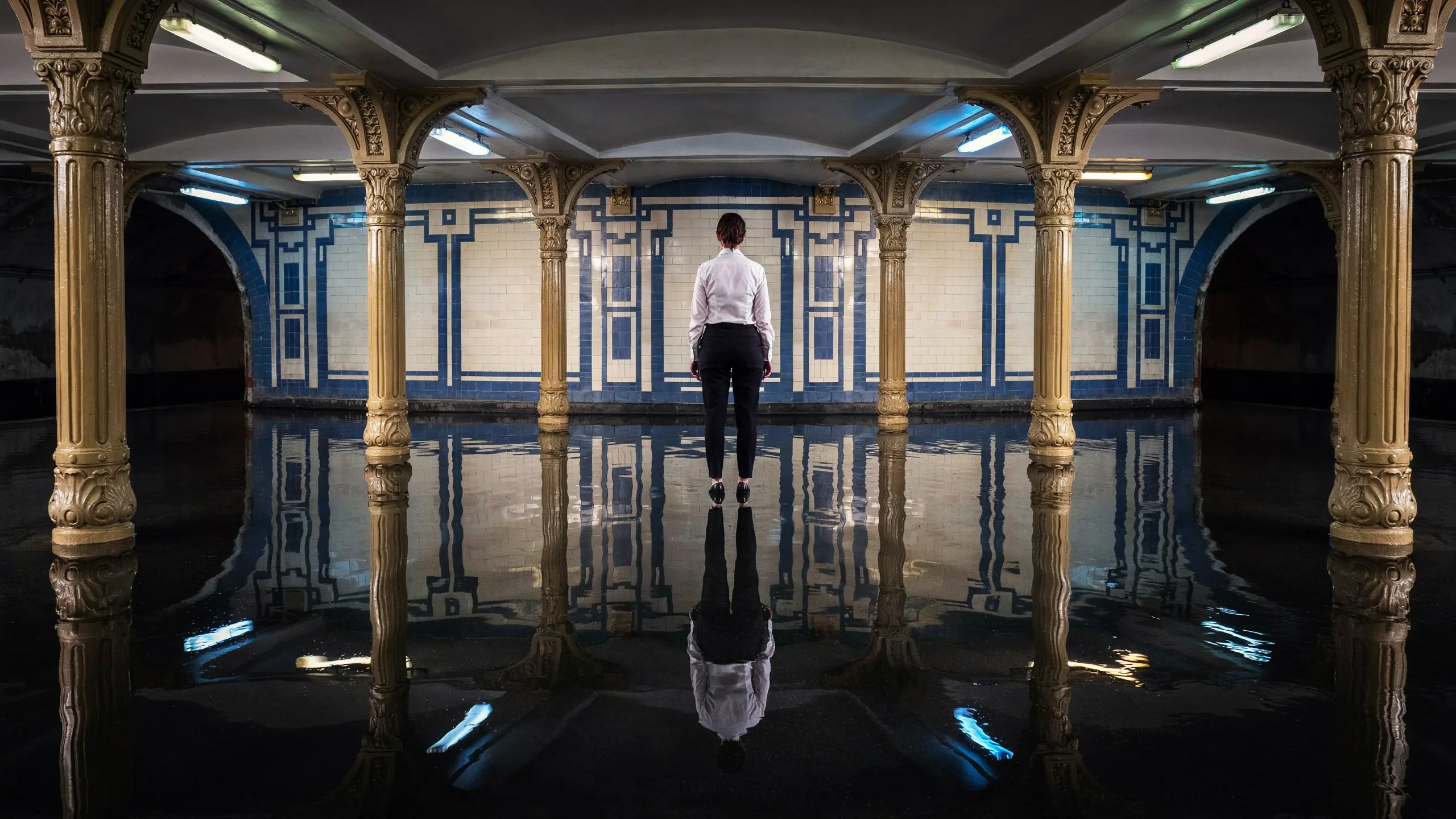 Flooded 1920s Art Deco underground station, a woman standing perfectly still in black water, back to the camera.