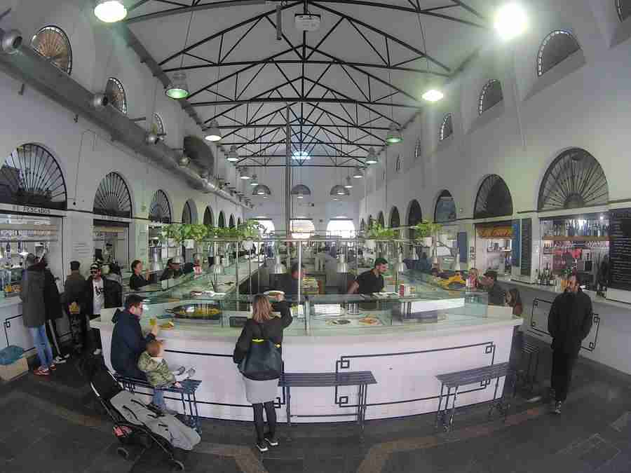 Vemos la galería de un mercado, con gente sentada comiendo en la barra y comprando alimentos a un lado.