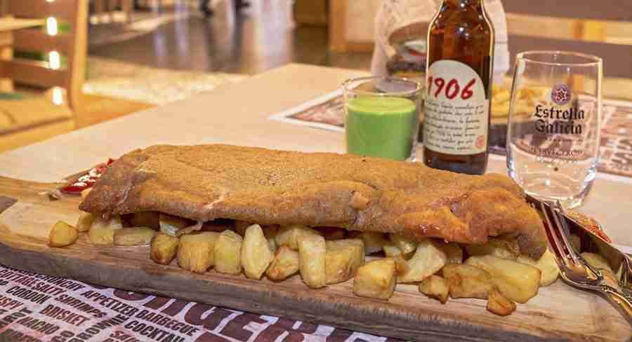 Un cachopo sobre patatas asadas, uno de los 5 Platos Fuertes de la Cocina Asturiana, sobre una tabla de madera y una cerveza de fondo