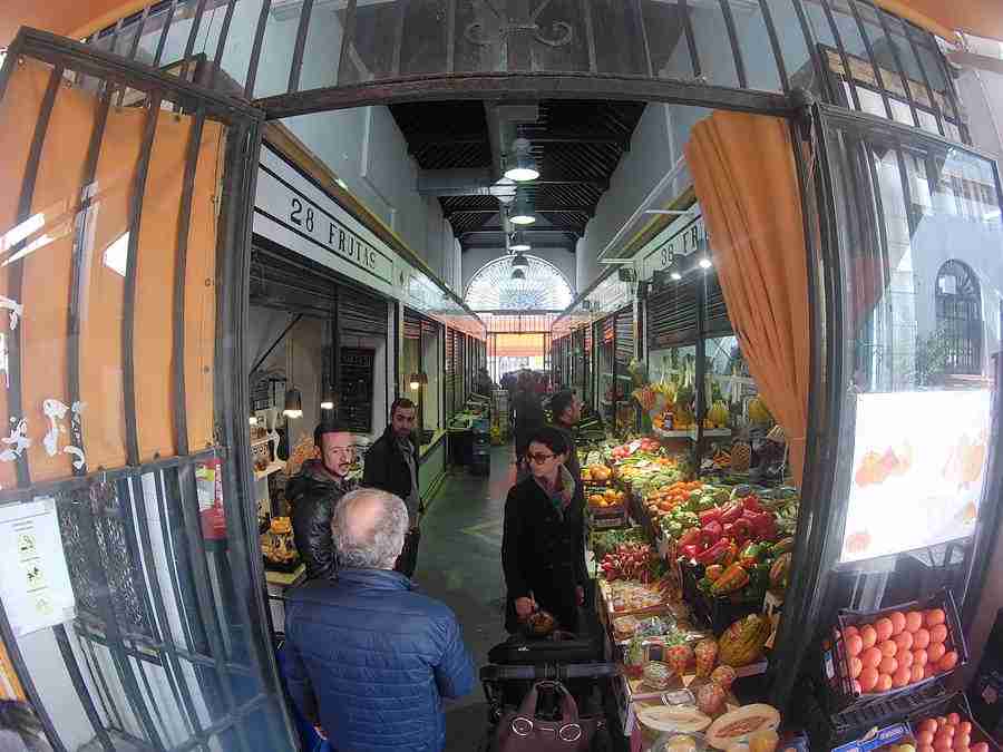 Vemos los pasillos de un mercado en Sevilla, con frutas y verduras exhibidas en un stand y gente comprando.