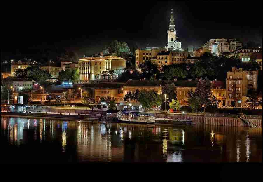Vemos la ciudad de Belgrado sobre el rio Danubio, a la noche, con una iglesia de fondo.